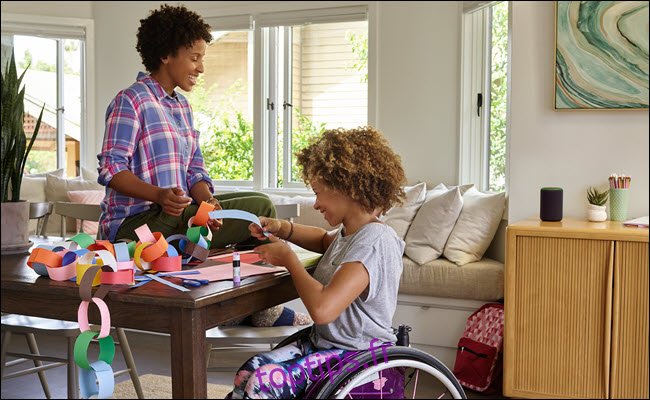 Un jeune enfant dans un fauteuil roulant faisant de l'artisanat avec sa mère à une table avec un Amazon Echo à proximité.