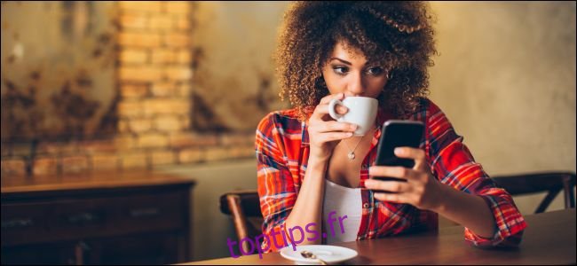 Une femme assise à une table en regardant un smartphone et en buvant du café.