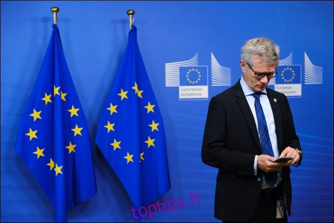 Un homme utilisant un smartphone avec deux drapeaux européens derrière lui.