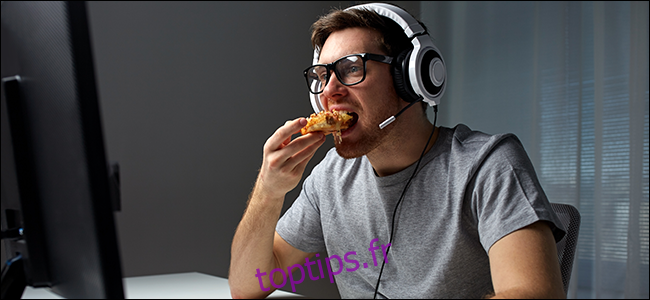 Un homme assis devant un ordinateur, portant un casque et mangeant de la pizza.